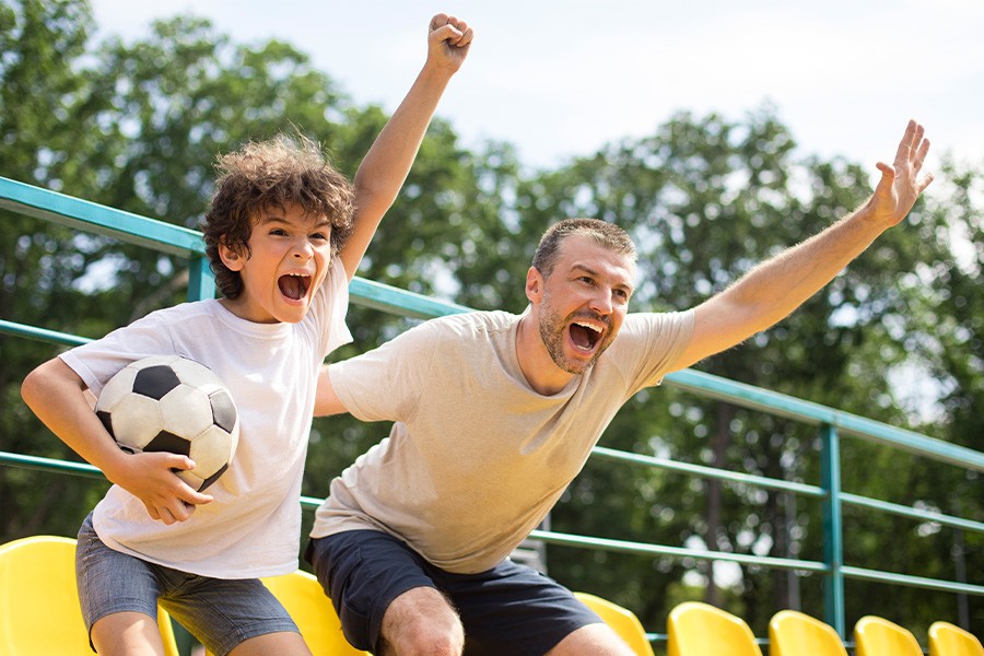 A man and a boy sit on a bench outdoors, arms raised in excitement, enjoying a fun moment together