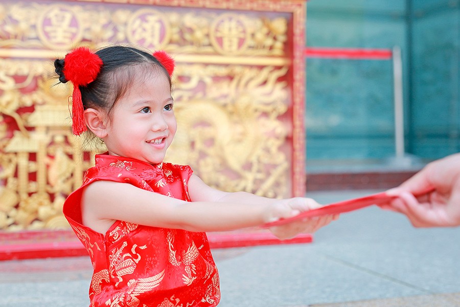 A little girl dressed in a vibrant Chinese outfit displays a red envelope, representing the Ang Bao tradition for New Year